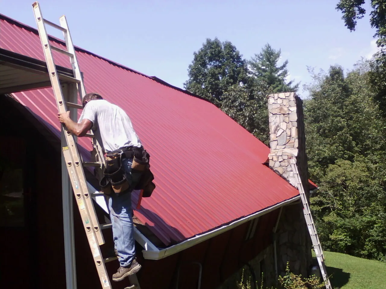 Roofer on a ladder beside a red metal roof and stone chimney — Soffit & Fascia Repair by Roofing Brothers in Palmersville